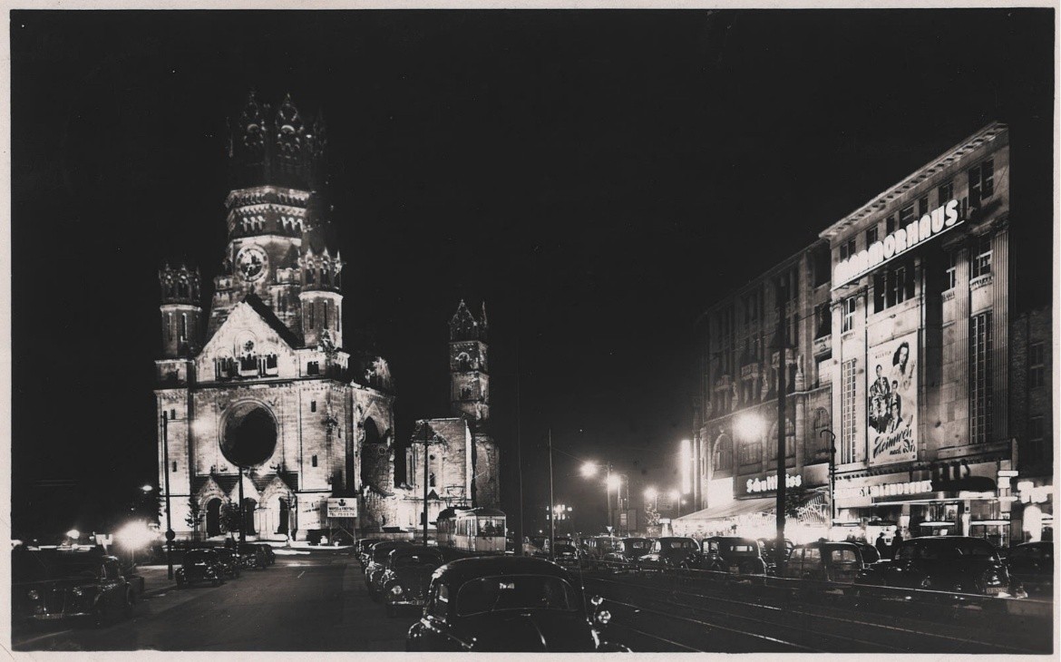 Night photograph of the Kurfuerstendamm: the floodlit Kaiser-Wilhelm-Gedaechtniskirche at left and, at right, the lit facade of the Marmorhaus carrying a large painted film poster, with cars parked the length of the street