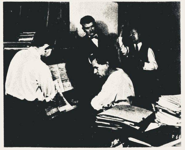 Grainy halftone photograph of four men around a table heaped with sheet music, one bent over sorting the scores while another leans across an open score on a stand