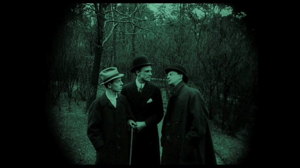 Green-tinted film still, pillarboxed in black. Three men stand together on a path in a bare wintry park among leafless trees. At the left a man in a light hat and dark overcoat looks to his right; in the centre a taller young man in a bowler hat and dark double-breasted coat holds a walking stick in front of him; at the right a man in a flat cap and dark overcoat stands in profile facing the other two