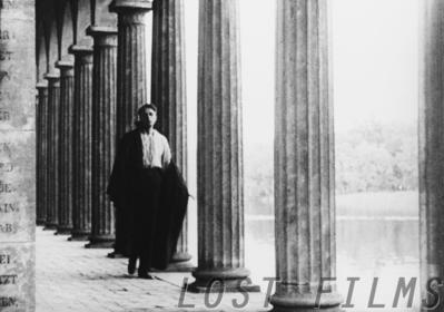 Film still: a man in a dark cloak stands among the tall columns of a colonnade, a lake visible through the gaps behind him