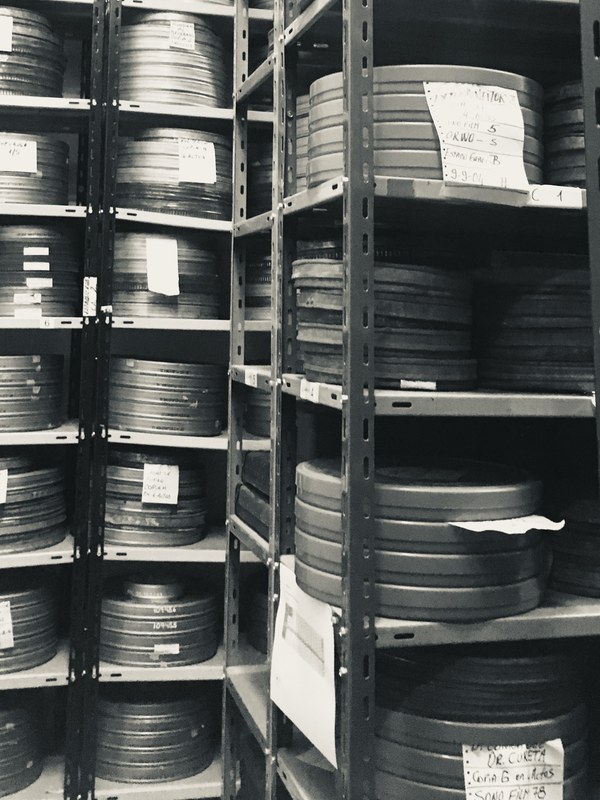 Metal film cans stacked on steel shelving in an archive vault, many carrying handwritten paper labels recording copy, reel and condition details